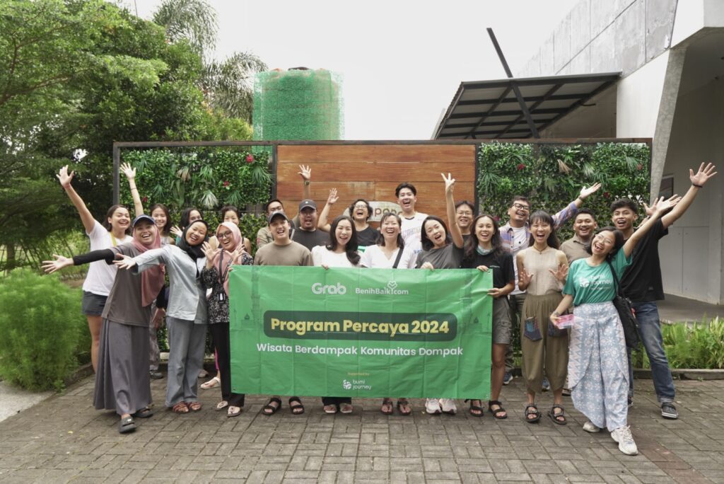 Kelompok peserta Program Percaya 2024 berpose ceria bersama sambil memegang spanduk bertuliskan "Wisata Berdampak Komunitas Dompak", di depan area hijau yang asri. Gambar ini menggambarkan contoh event organizer yang sukses menyelenggarakan acara wisata berdampak positif.