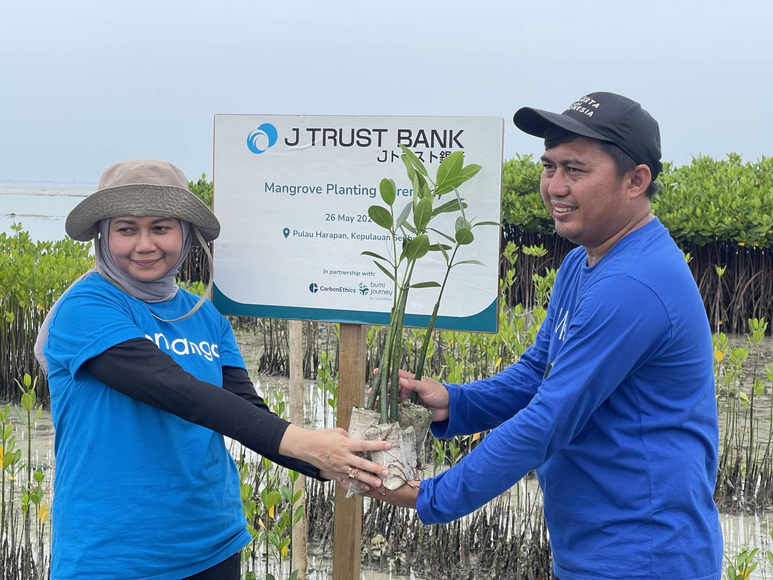 Perwakilan pihak J Trust Bank memegang bibit-bibit mangrove bersama perwakilan petani mangrove secara simbolis di depan papan plakat nama J Trust Bank