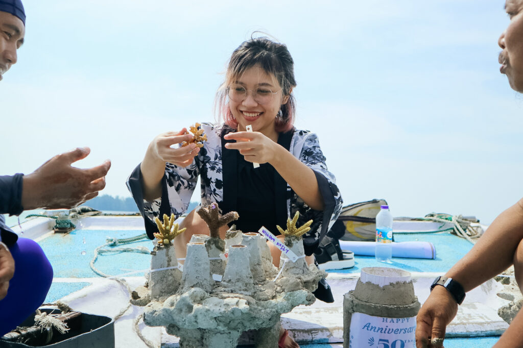 wanita berkacamata memegang bibit koral di atas perahu di temani staf pengurus lokal pria
