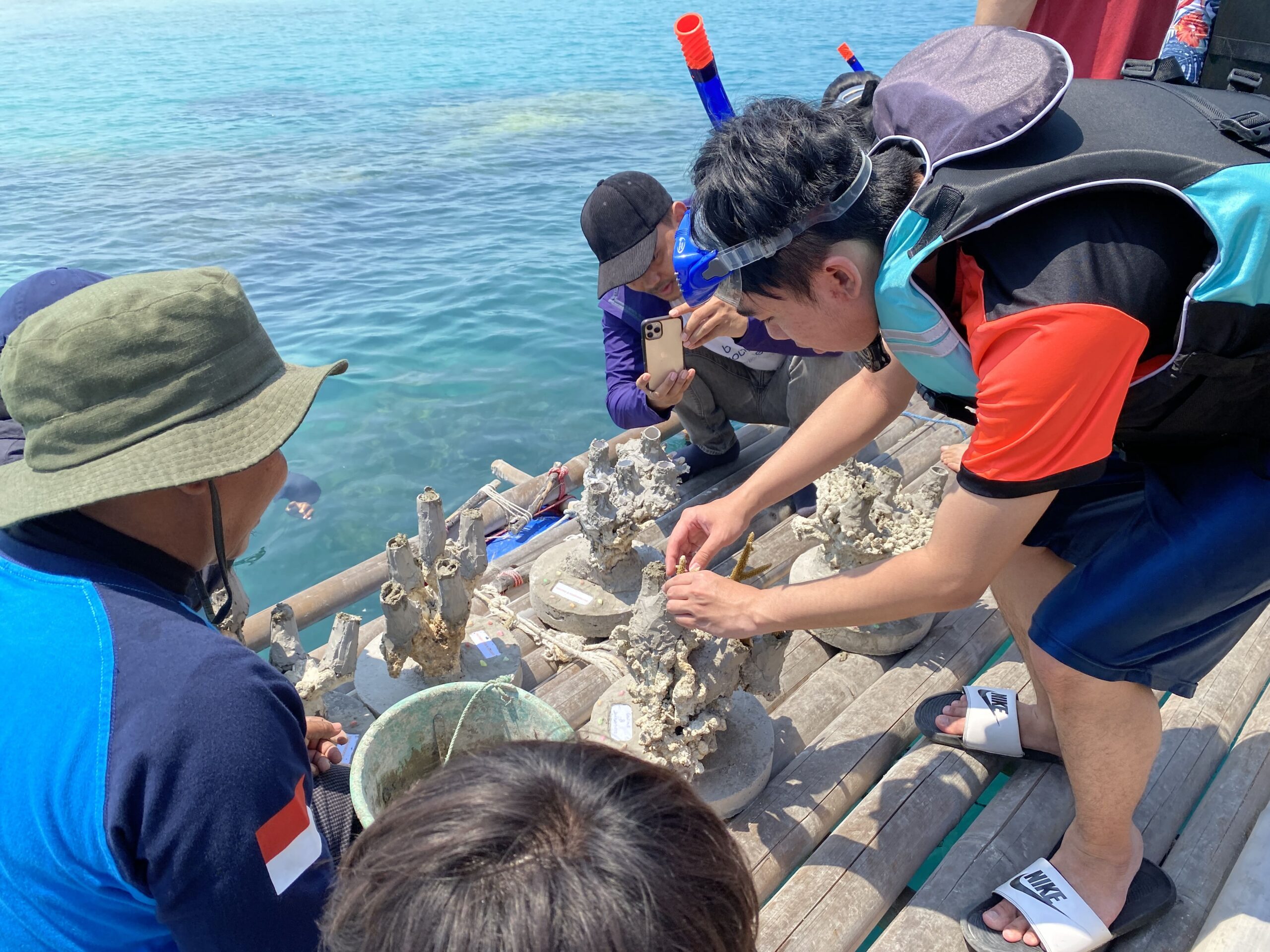 A man in snorkeling gear leaning down to affix a baby coral on the coral base surrounded by coral restoration projects workers on a wooden platform on the beach