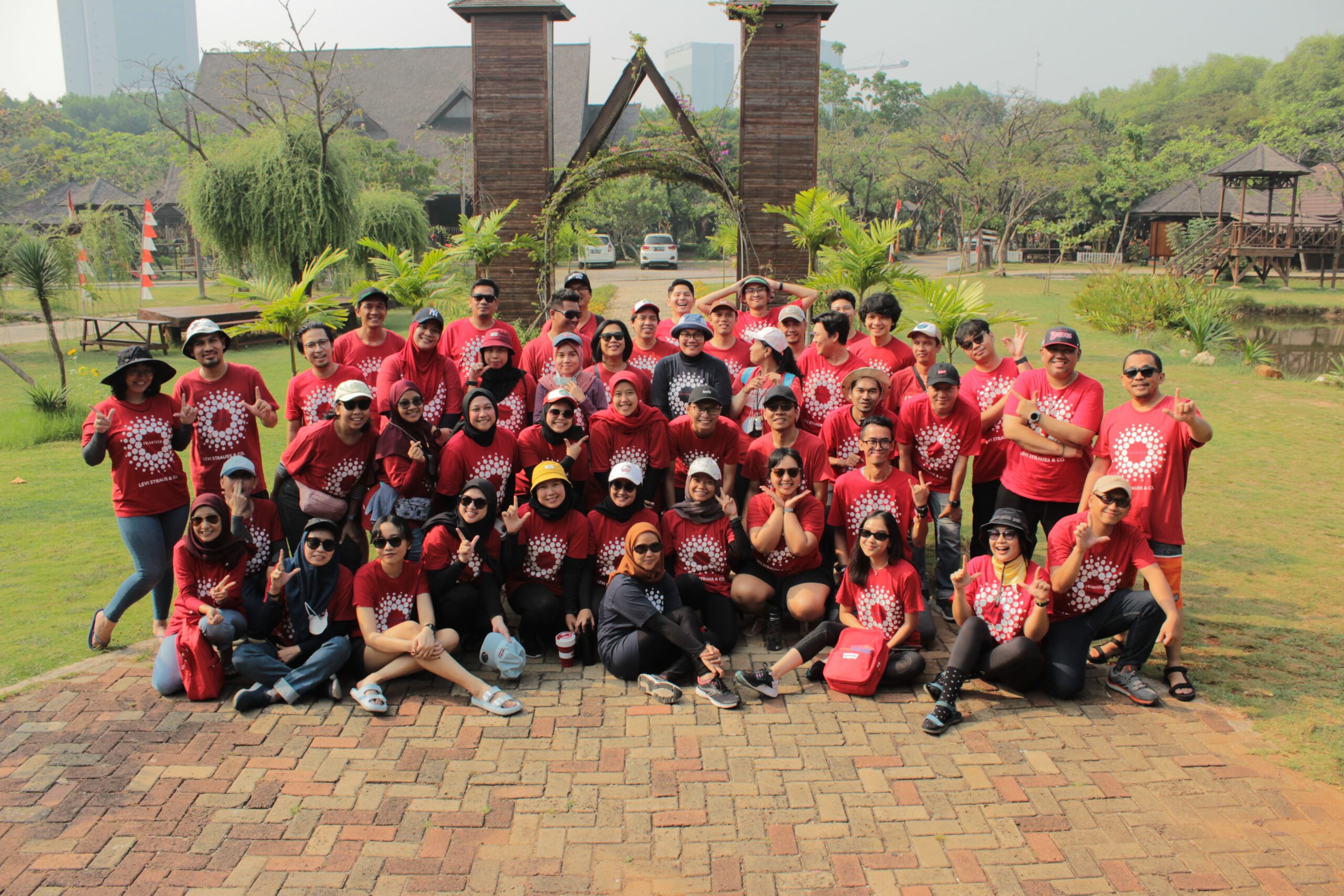 Foto kelompok besar peserta kegiatan outdoor mengenakan kaos merah seragam, berpose bersama di area taman dengan latar belakang hijau dan struktur kayu. Ini adalah contoh event organizer dalam mengelola acara team building untuk perusahaan.