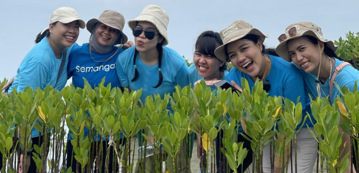 6 wanita berbaju biru Bumi Journey dengan bucket hat sedang gathering kantor berdiri di belakang bibit mangrove di pesisir pantai