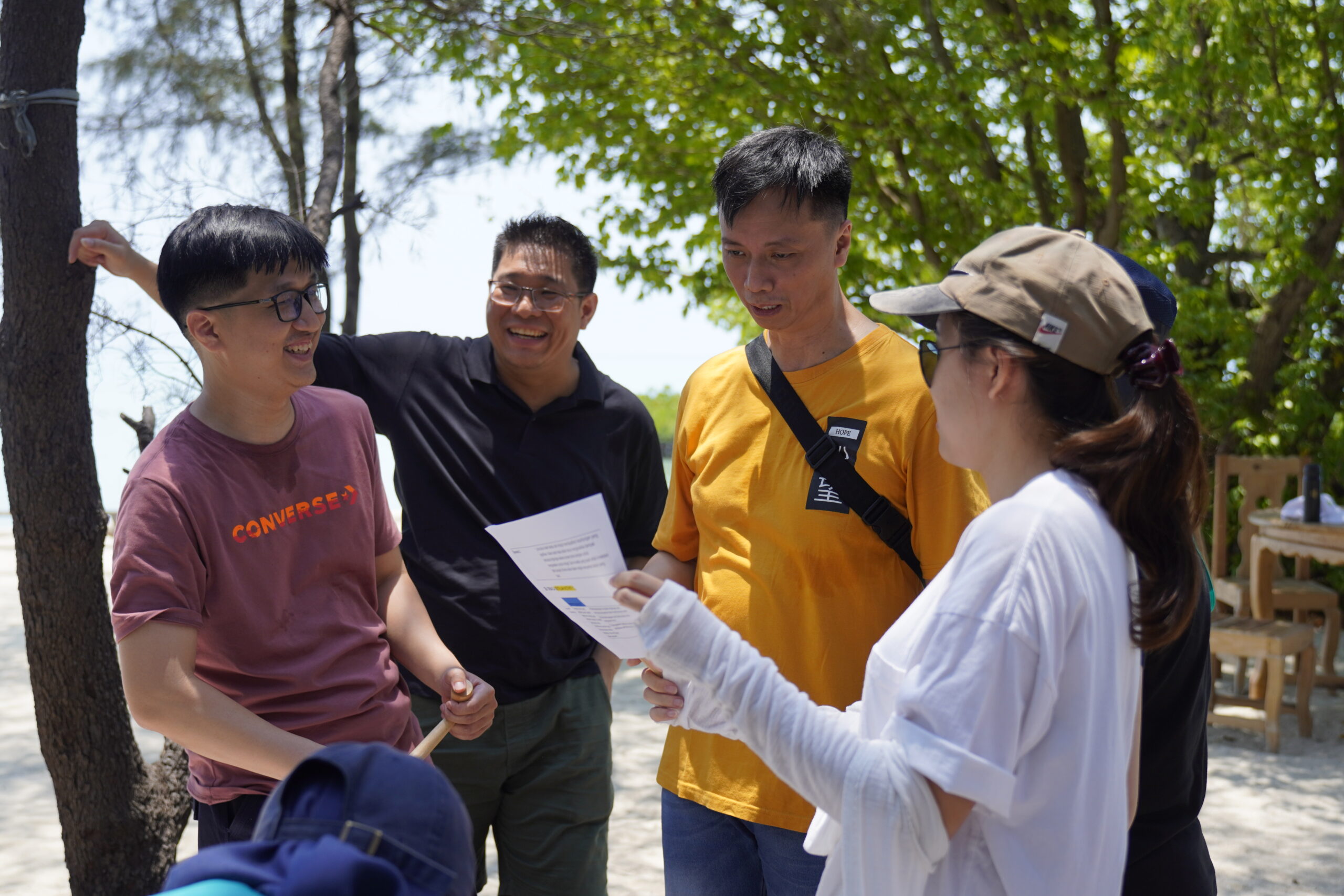 3 pemuda dan 2 pemudi tersenyum gembira sambil bertukar pikiran (brainstorming) di tepi pantai dalam acara gathering kantor