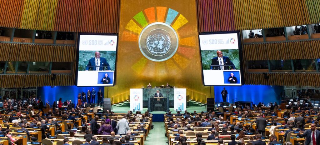 United Nations SDG Summit in a giant conference room with a black male speaking on the podium displayed on two screens to the left and right