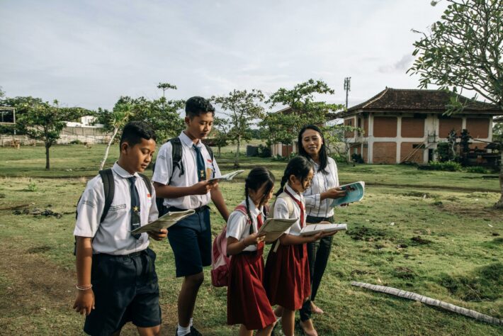 2 anak perempuan SD dan dua lelaki SMP berseragam belajar di ruang terbuka suasana pinggiran kota hijau bersama seorang guru wanita sambil memegang buku